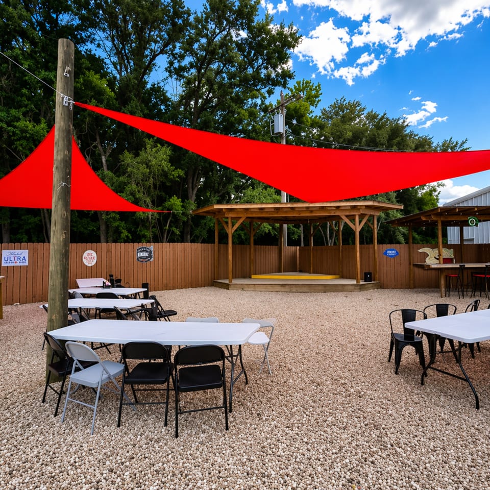 Outdoor dining area with white tables, black chairs, red shade sails, and a wooden stage.