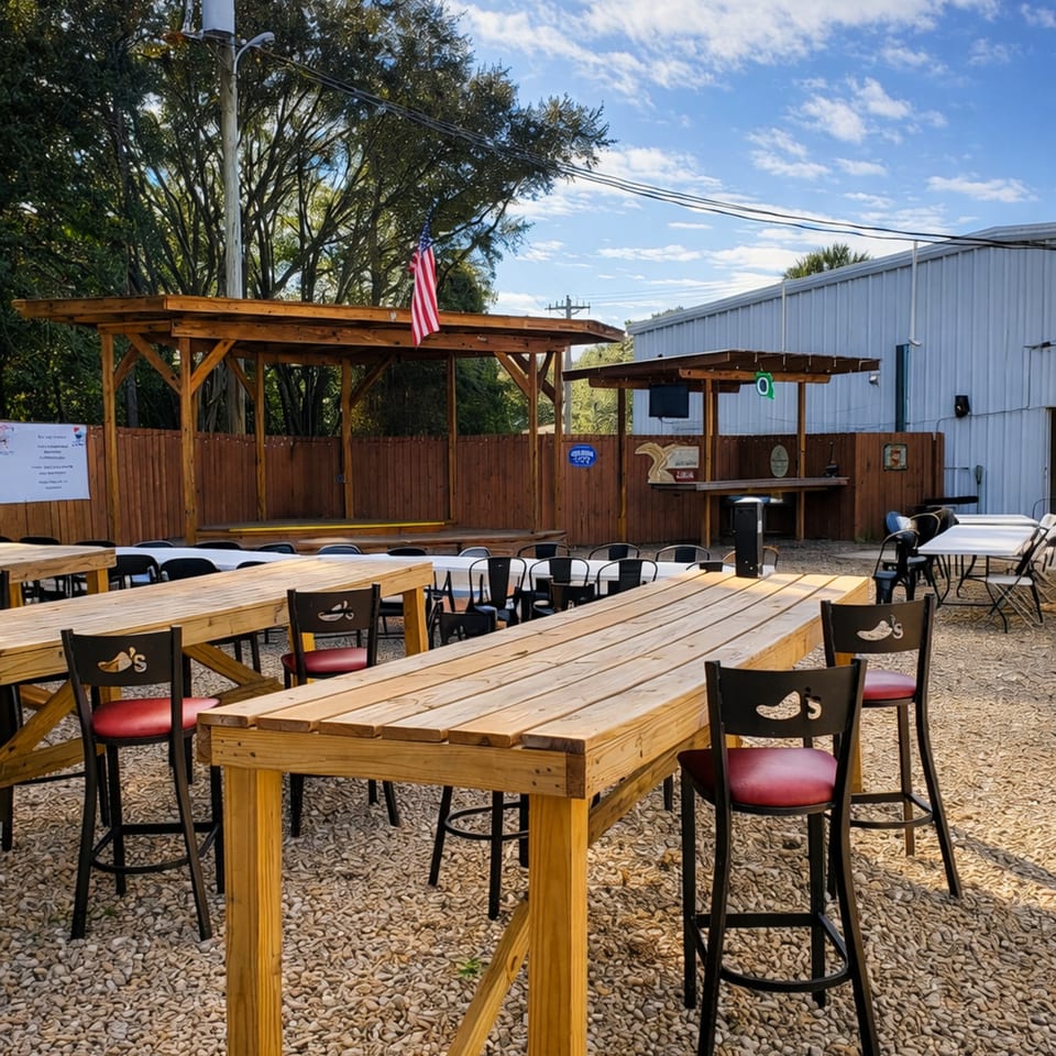 Outdoor dining area with wooden tables, red-seated bar stools, and a covered stage under a blue sky.